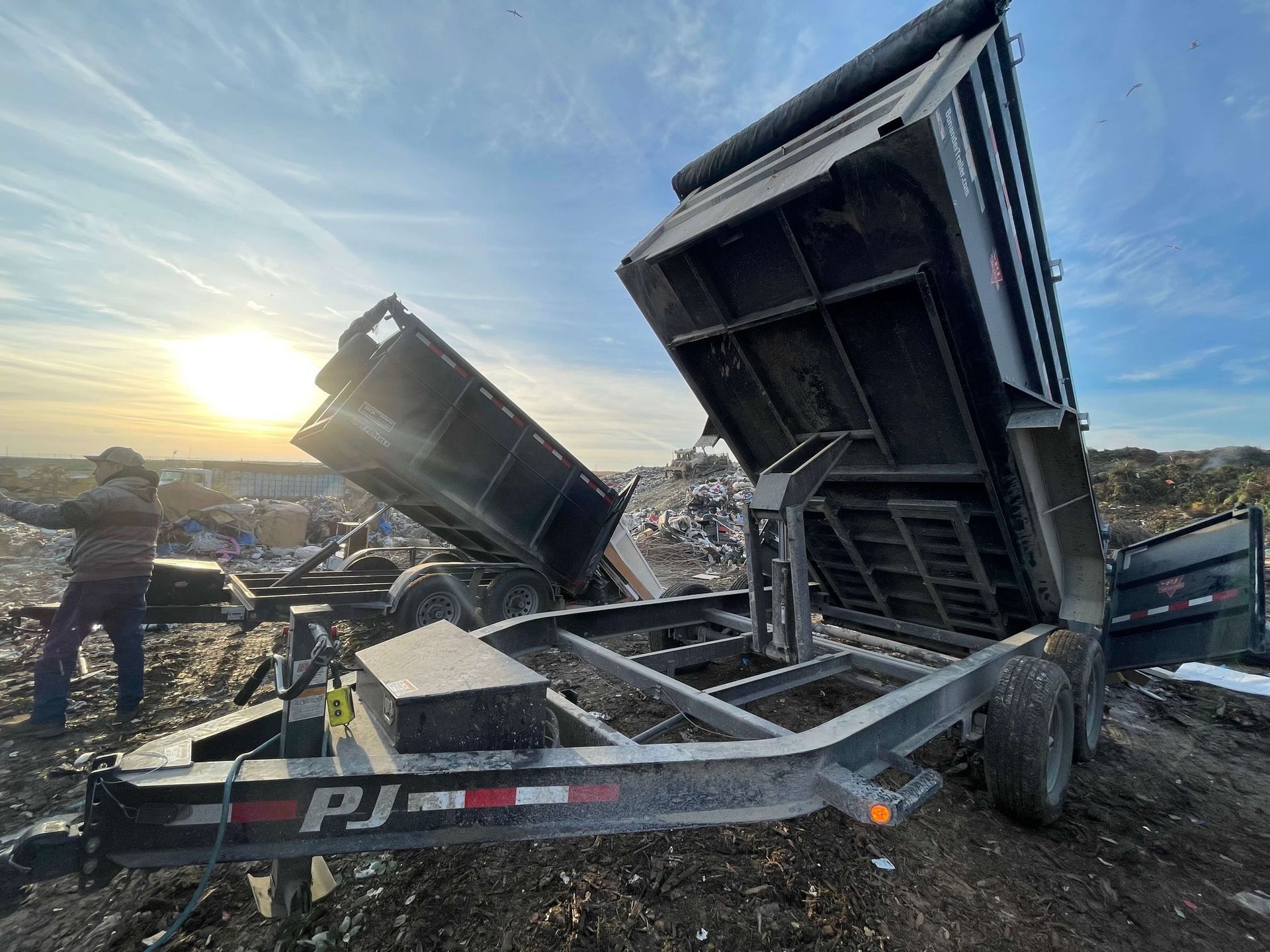 Two PJ brand dump trailers lifted in a field under a sunset sky, with a person standing nearby.
