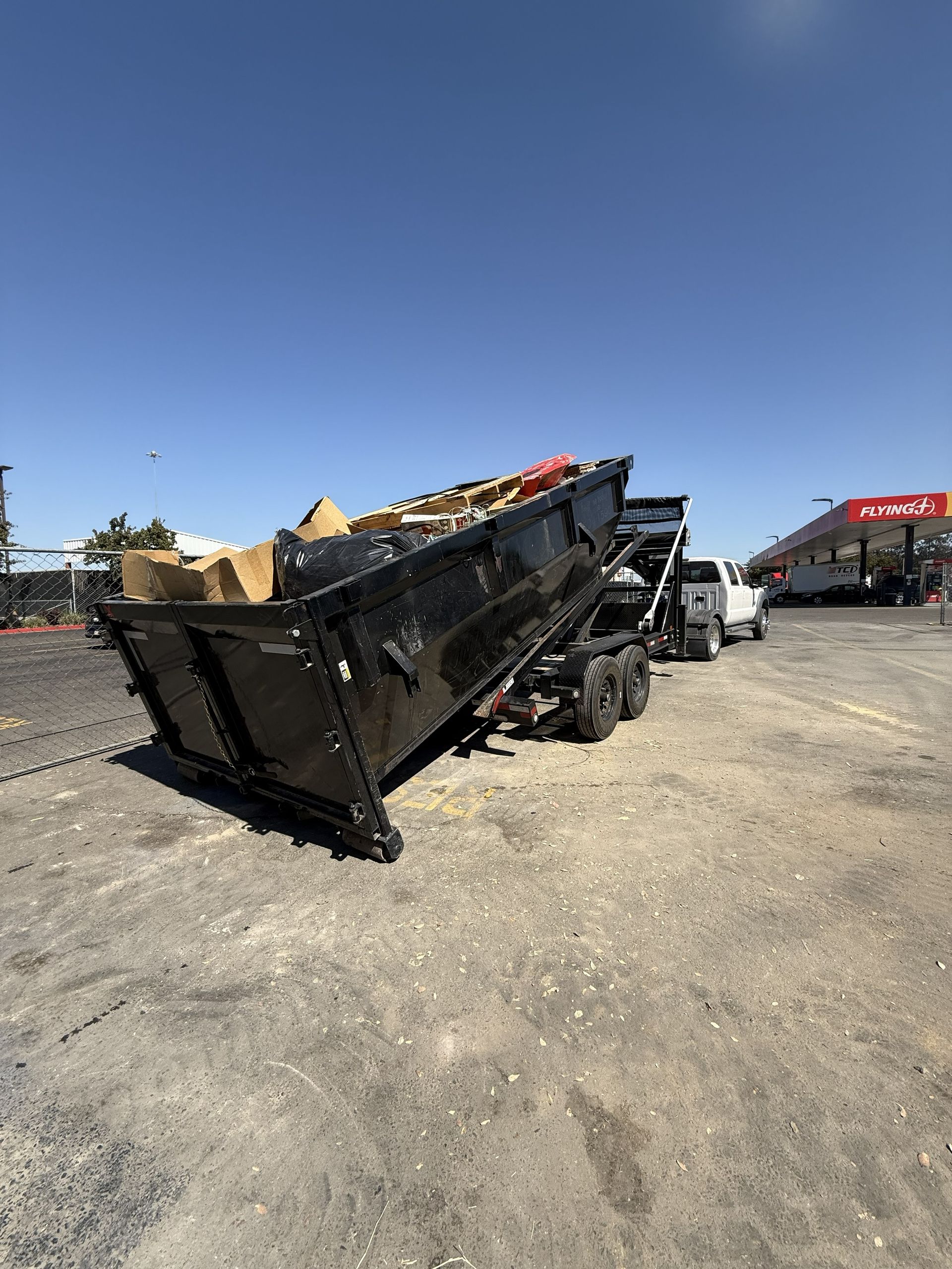 A black dump trailer loaded with construction debris sits on a gravel lot near a gas station under a clear blue sky.