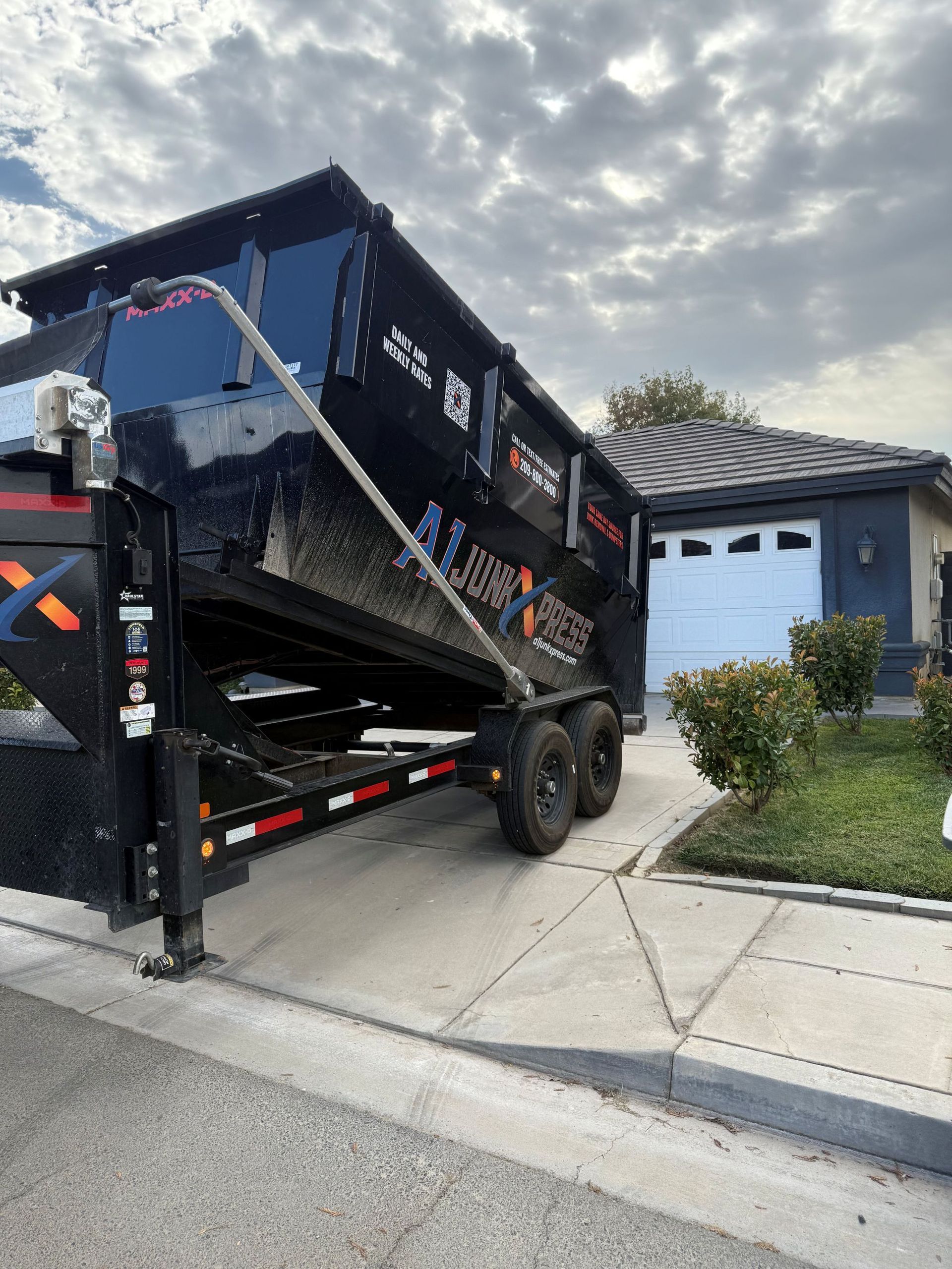 A large, black dump trailer parked in the driveway of a residential house on a cloudy day.