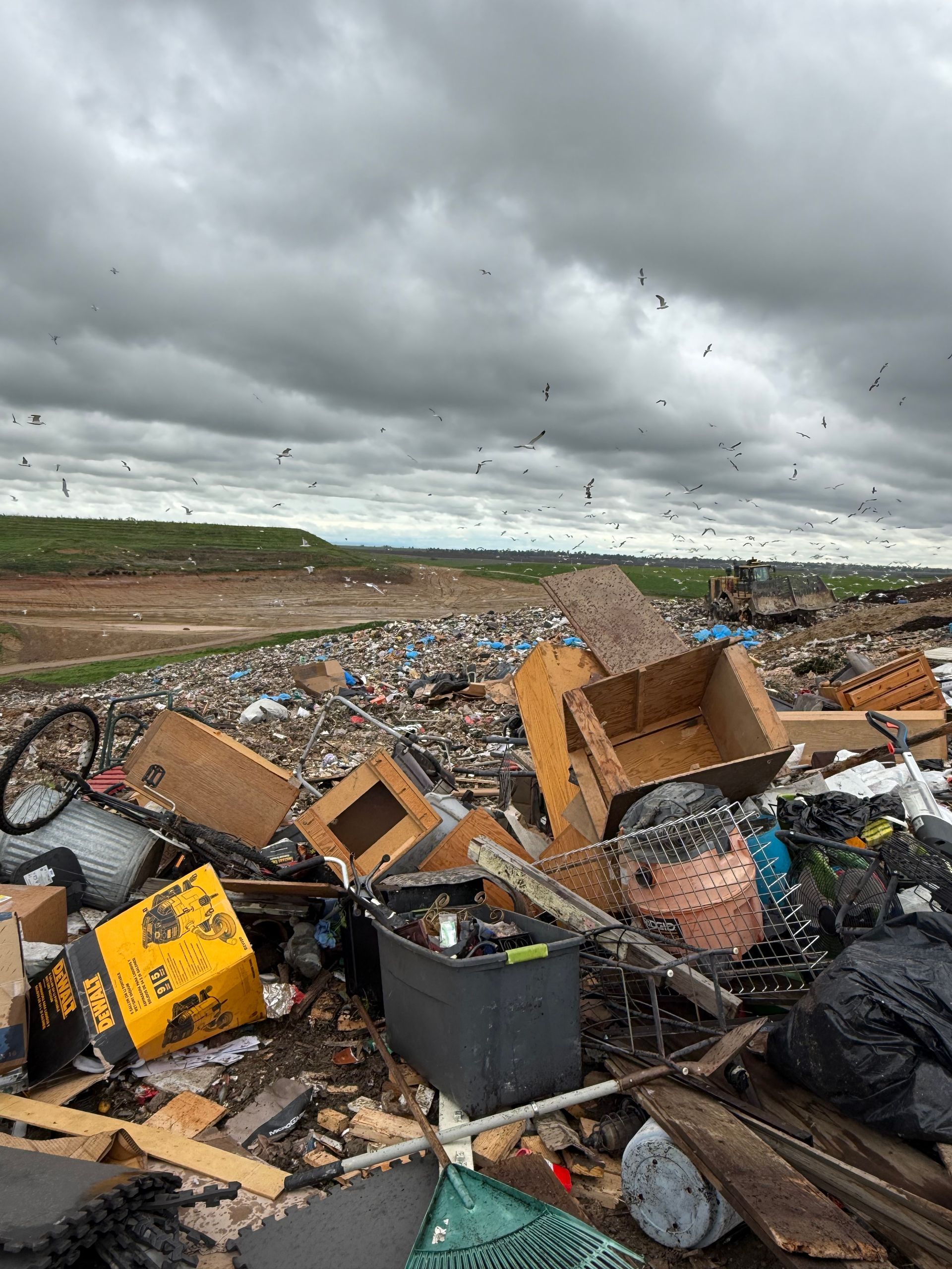 A wide, elevated view of a large, messy landfill under a dark, cloudy sky, with many birds flying overhead.