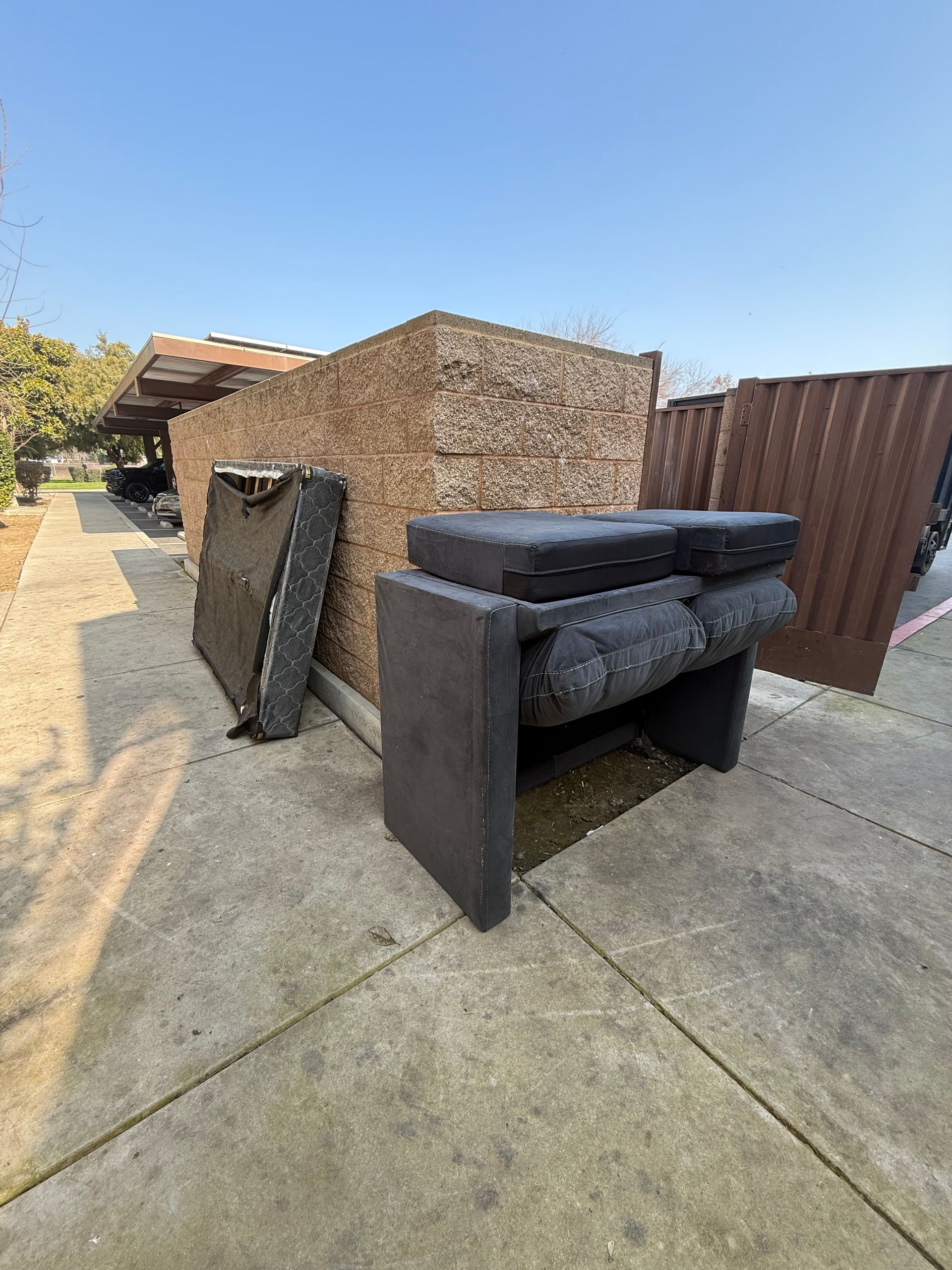 A stack of dark gray cushioned furniture and a mattress leaning against a large wooden crate on an outdoor concrete path.