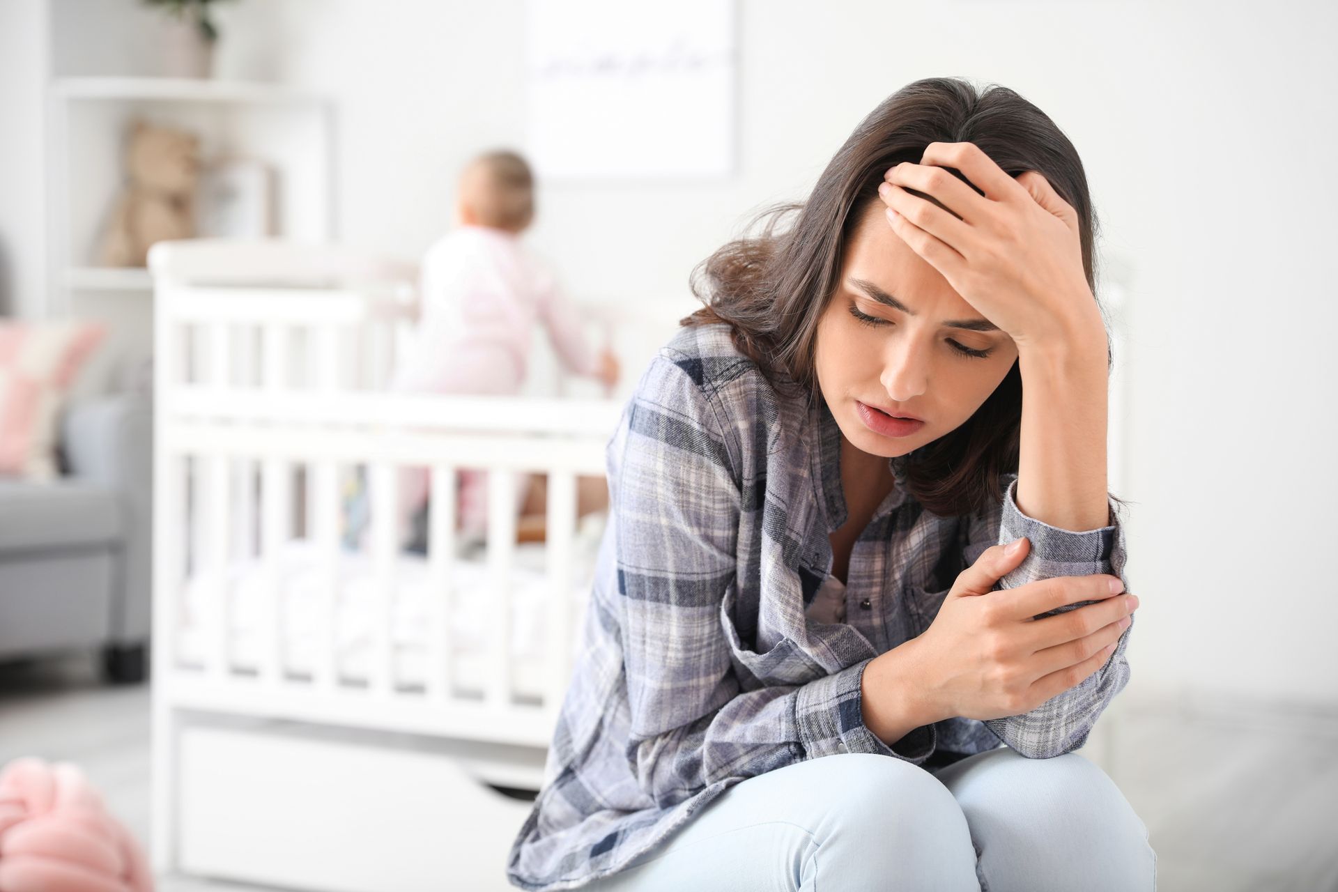 Woman sitting on the floor, holding her head, looking upset in a bright nursery with a baby in a crib