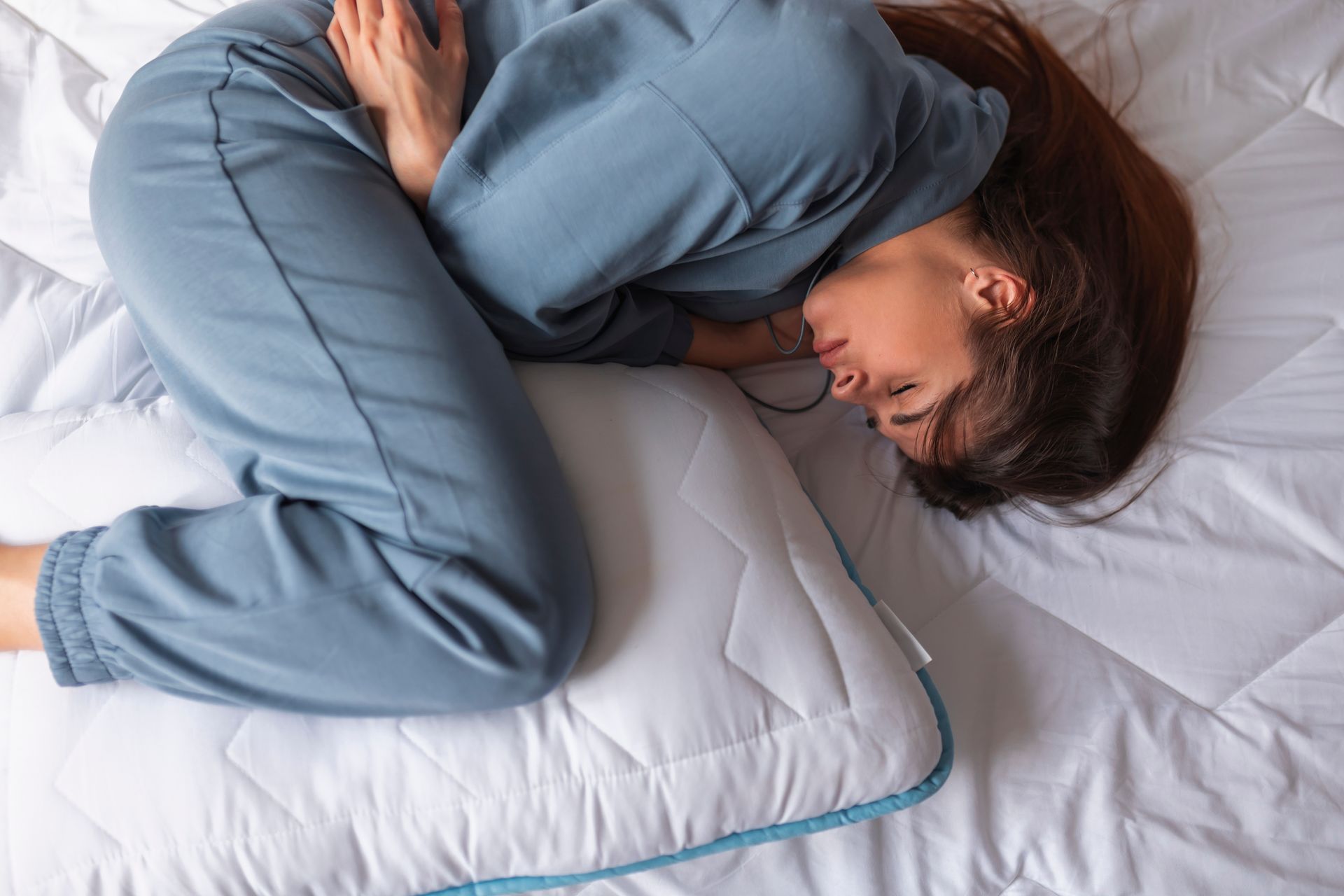 Person curled up on a bed with a hand on their head, appearing uncomfortable or in pain