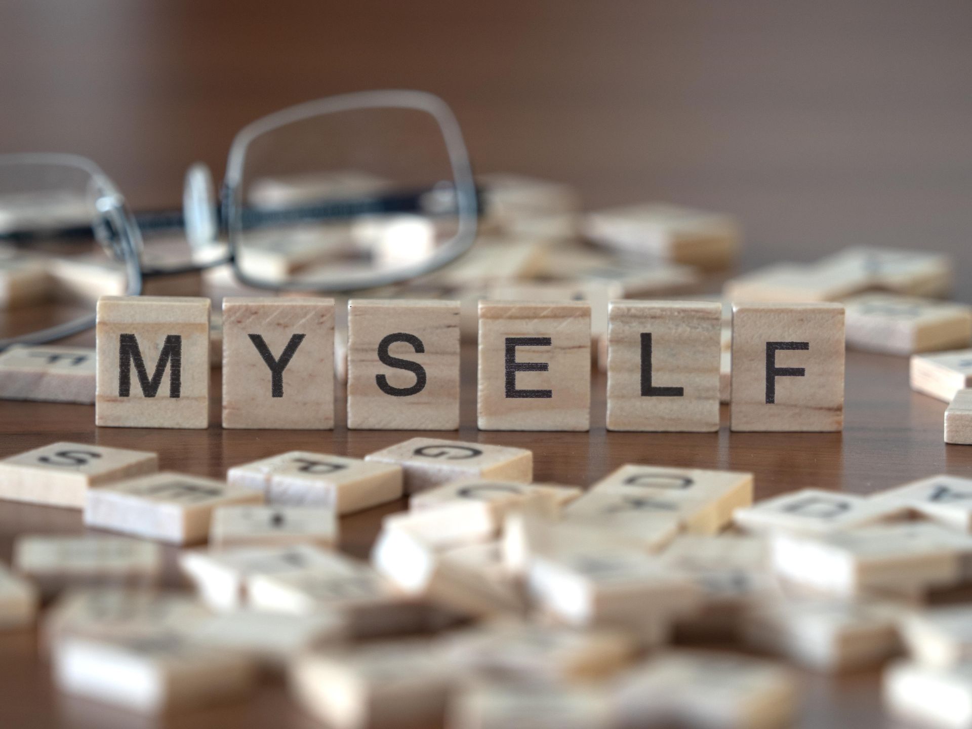 Wooden letter tiles spelling “MYSELF” among scattered tiles, with eyeglasses in the background.