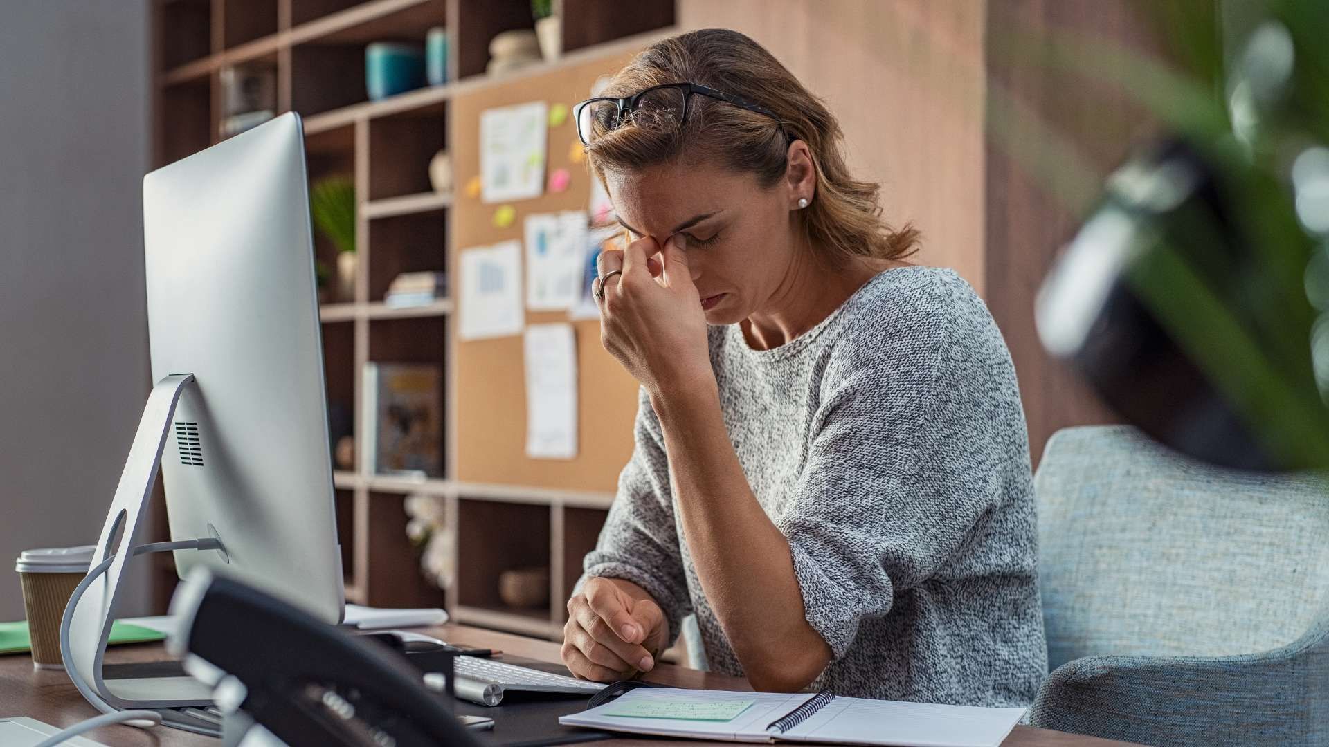Woman at a desk rubbing her eyes in an office, looking stressed beside a computer.
