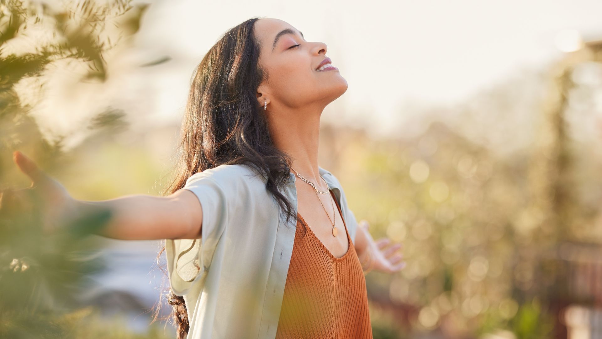 Woman with arms outstretched, smiling in a sunlit field with blurred flowers.