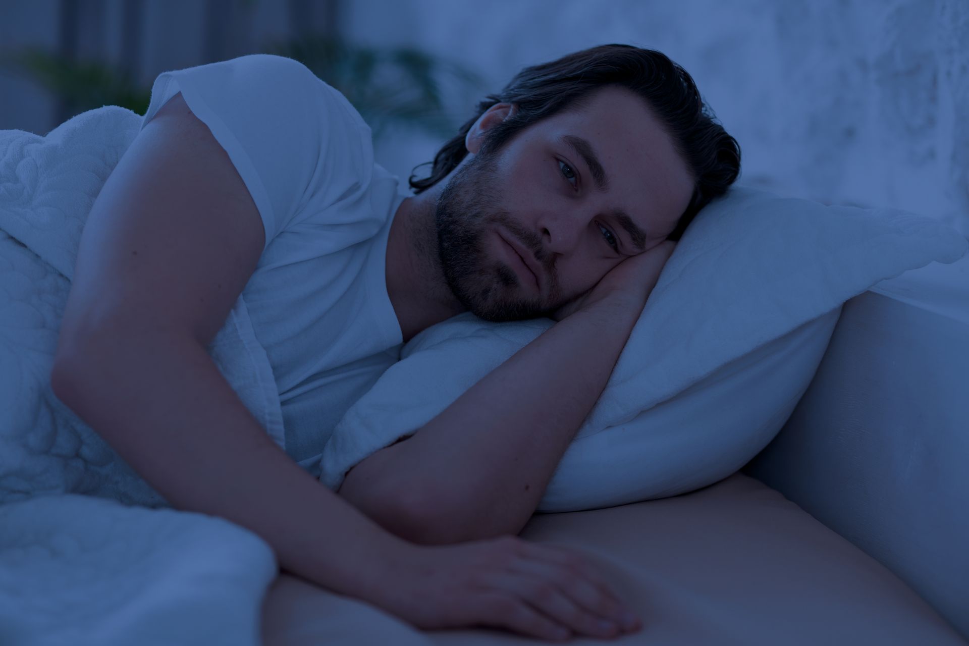 A man lying on a bed with a white pillow and blanket in dim blue light, looking tired or sleepy