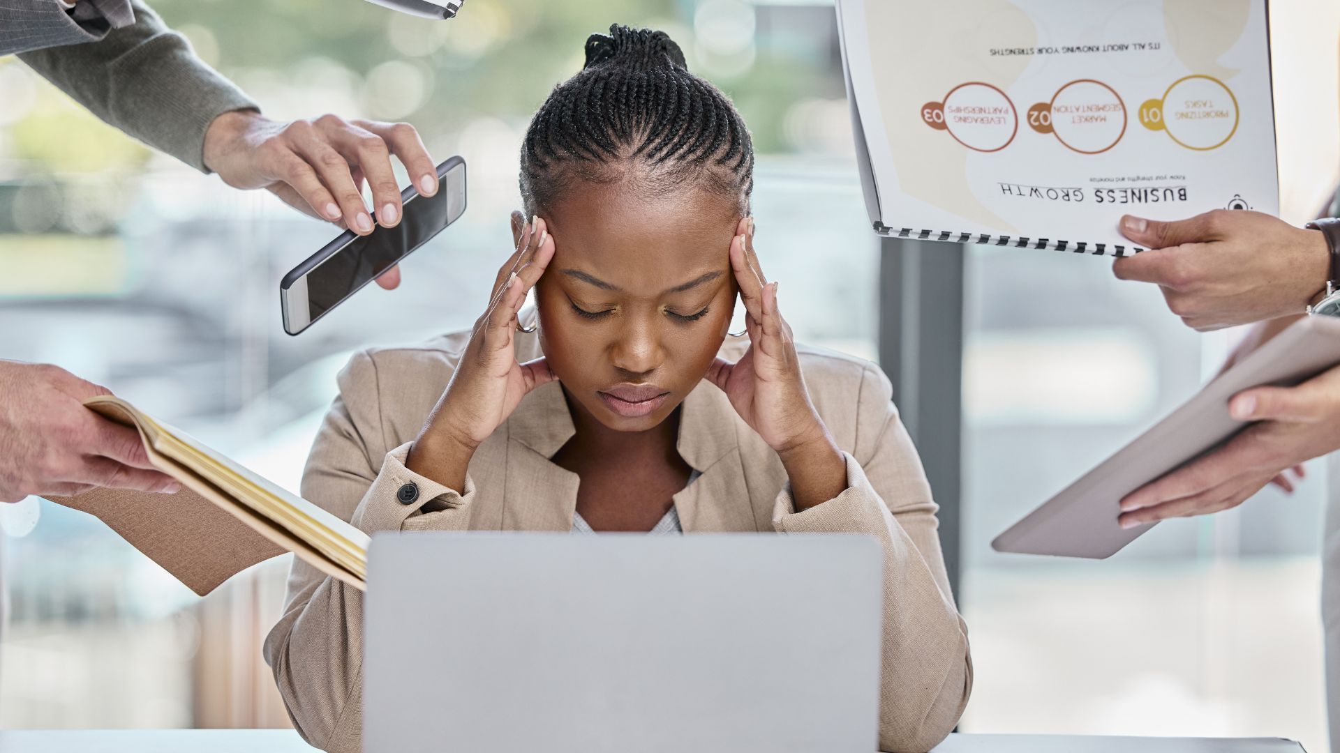 Stressed person at laptop with hands on temples, surrounded by papers and coworkers in an office setting