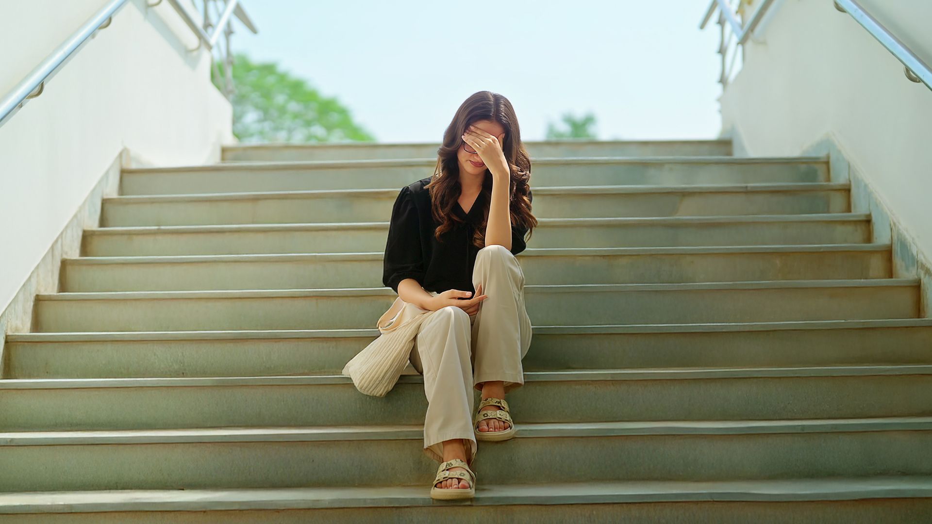 Person sitting alone on outdoor stairs, head in hand, with a white bag beside them.