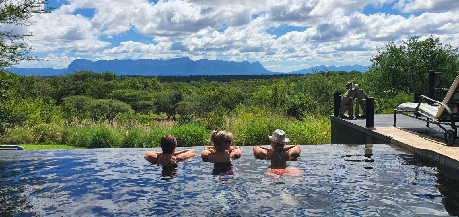 Three people are swimming in an infinity pool with a mountain in the background.