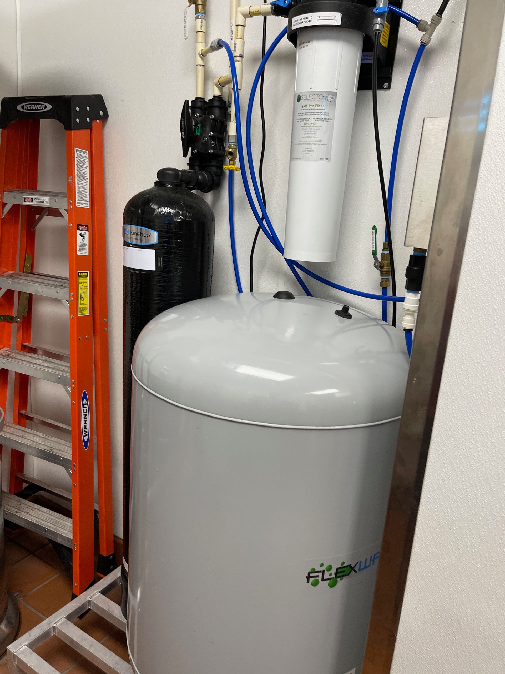 Water softener system with large white tank, black cylinder, and wall-mounted pipes in a utility room