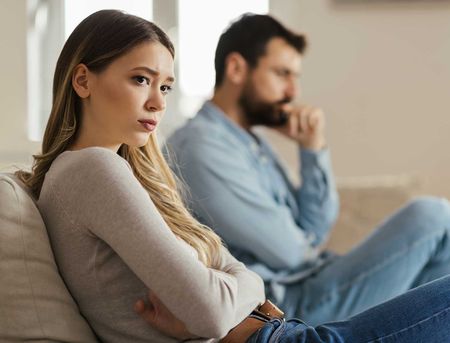 Worried young woman sitting on sofa at home and ignoring her boyfriend who is sitting next to her
