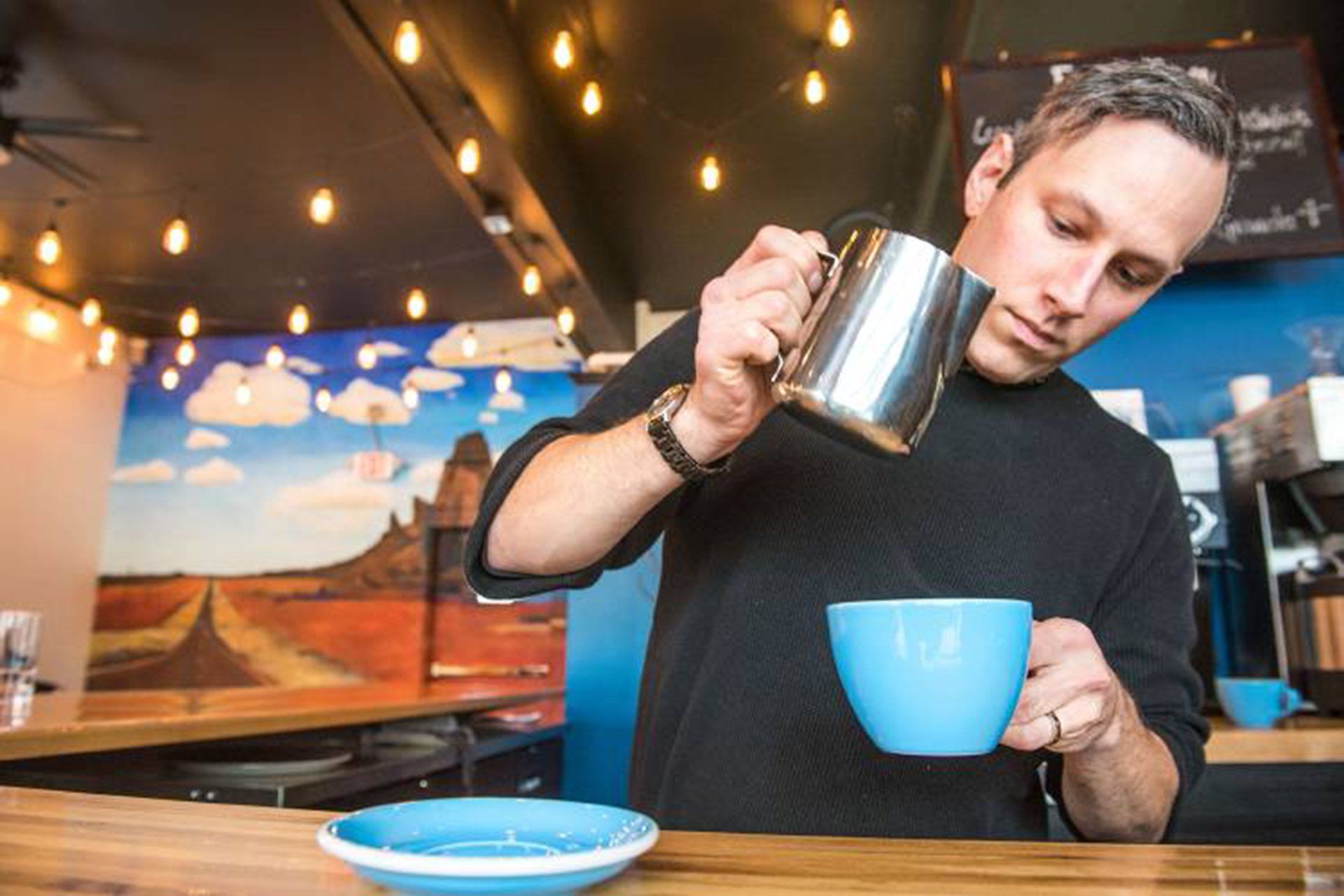 A man is pouring milk into a blue cup of coffee.