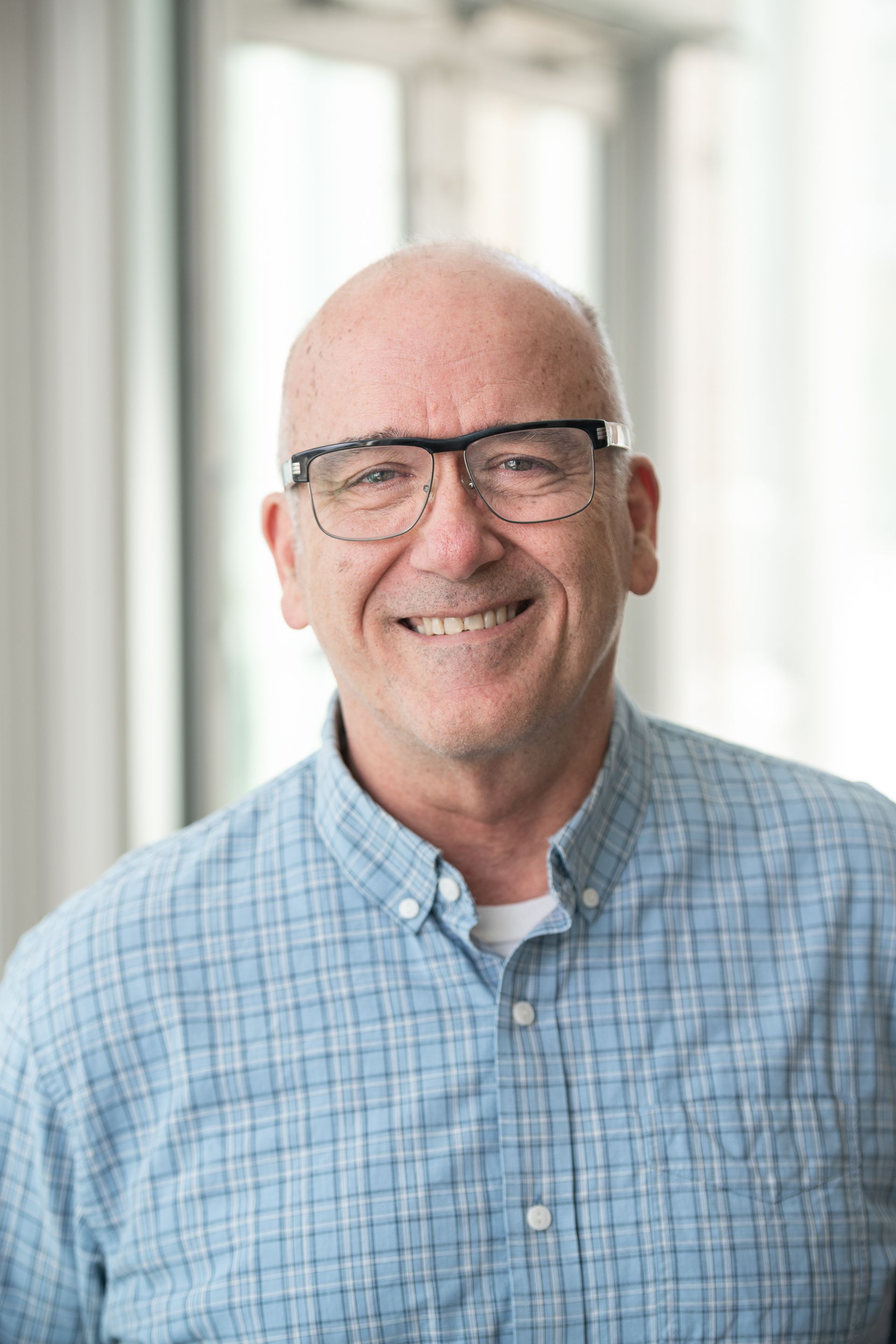 A bald man wearing glasses and a blue plaid shirt is smiling for the camera.