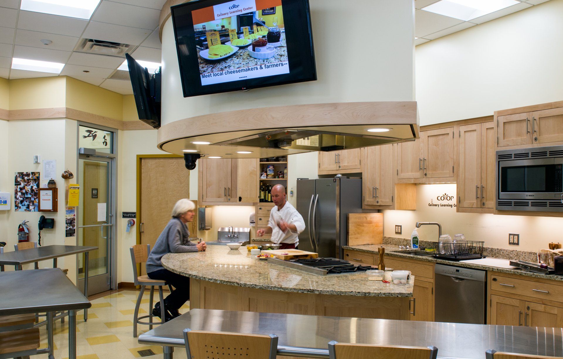 Two people are sitting at a table in a kitchen with a large flat screen tv above them