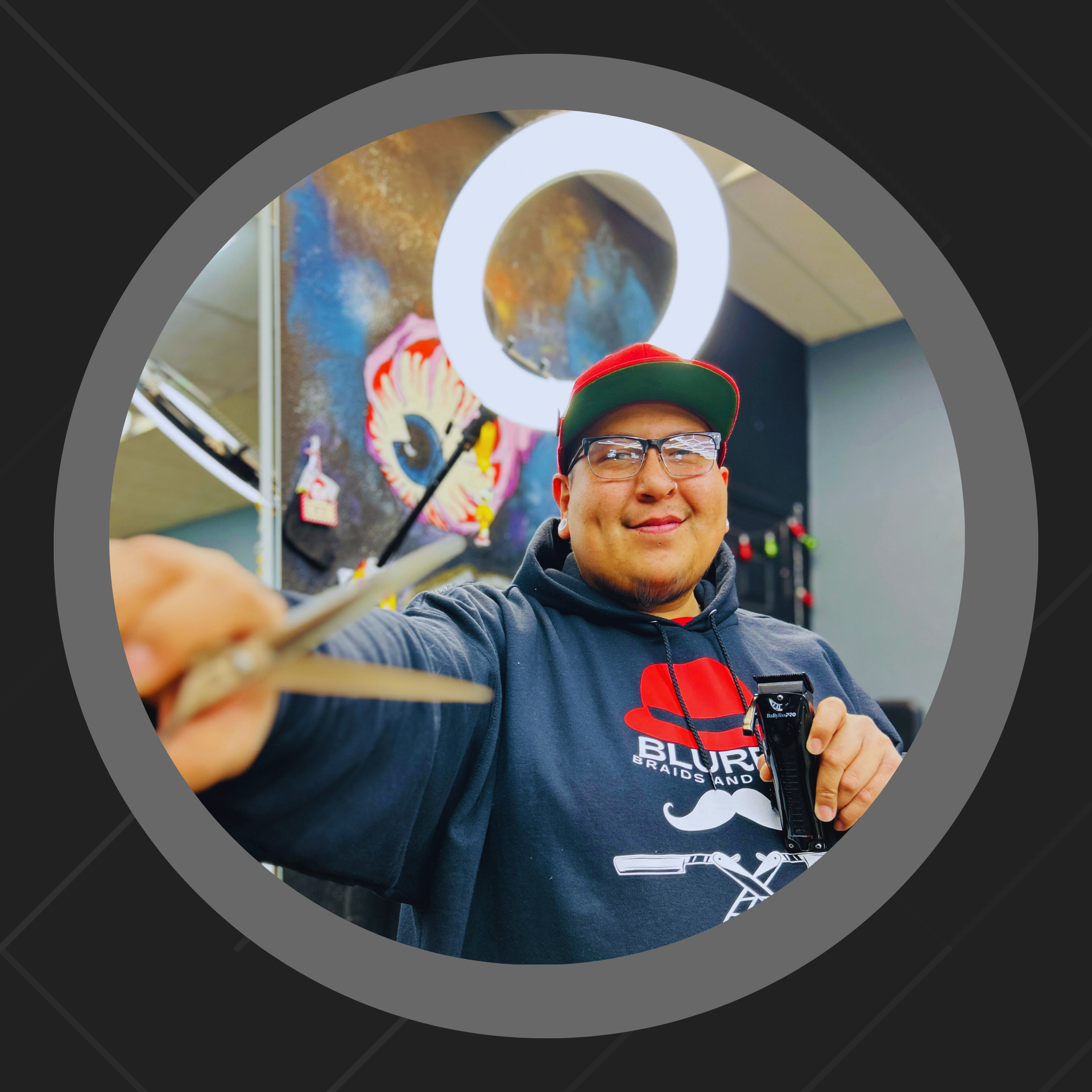 Barber holding scissors and clippers, wearing a hat and hoodie, indoors with artwork and a ring light.