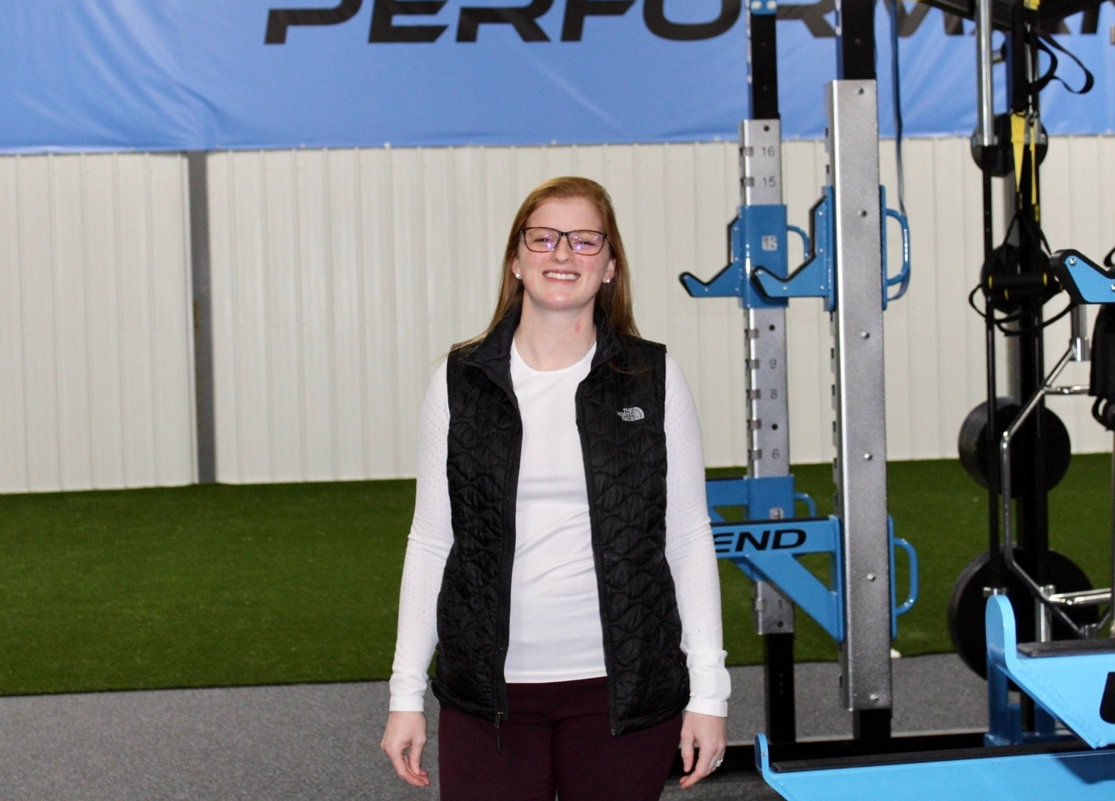 Woman in a black vest smiles in a gym, near exercise equipment with a blue accent.