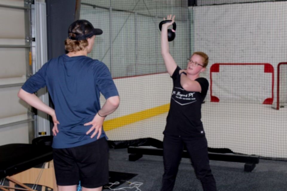 A person lifting a kettlebell overhead while being observed by another in a gym setting.
