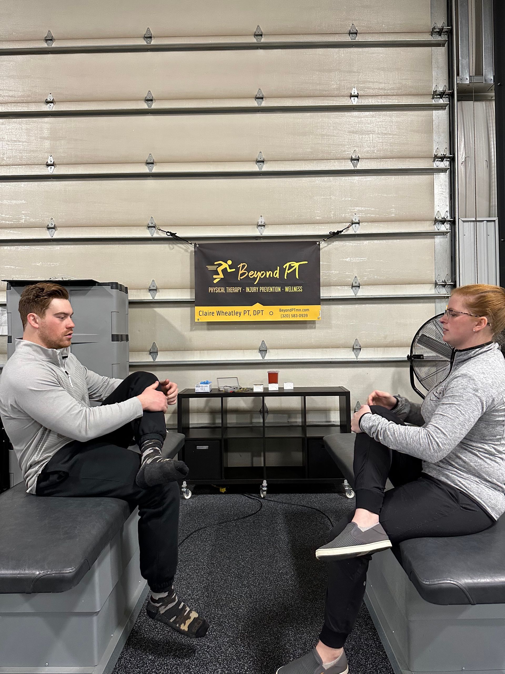 Two people seated on benches in a gym, legs crossed, facing each other in conversation.
