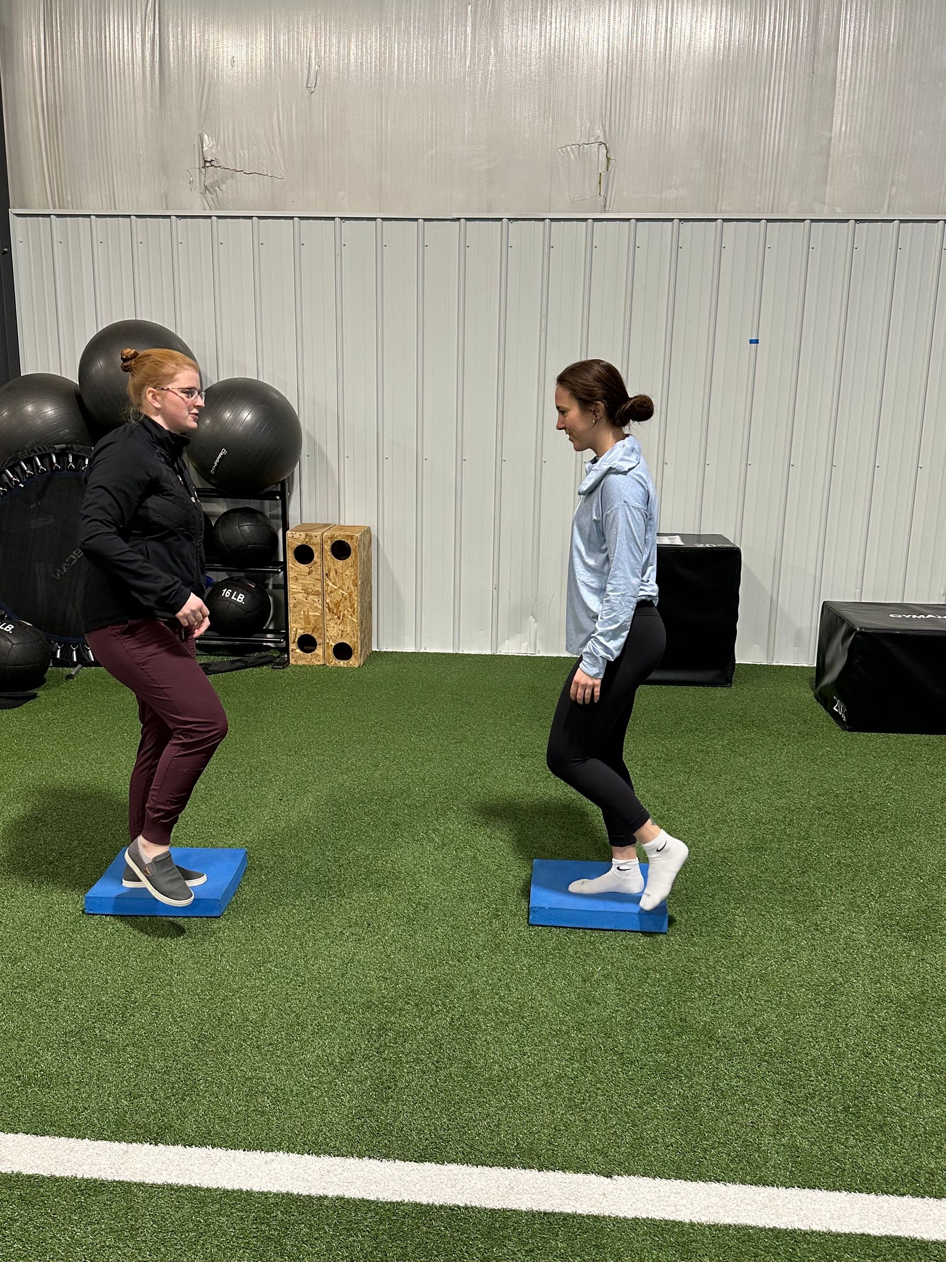 Two people stand on blue platforms on a green floor. They are performing exercises indoors.