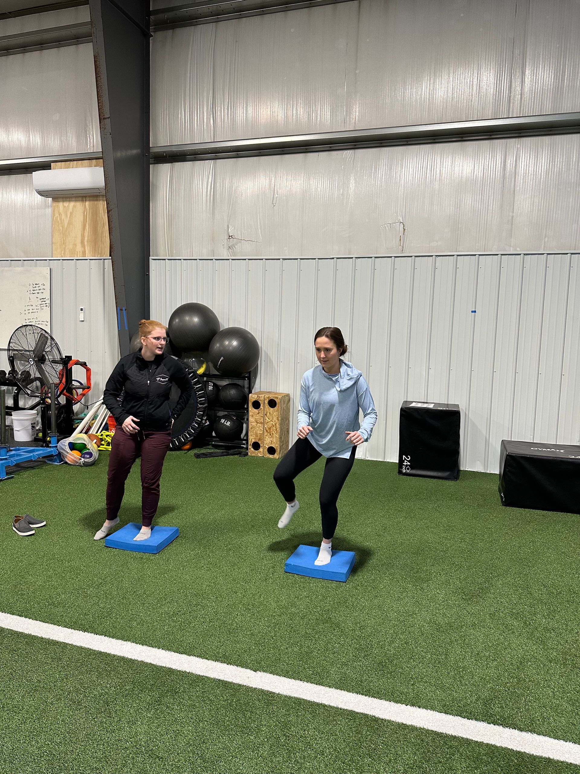 Two people exercising on blue mats in an indoor gym with equipment and turf.