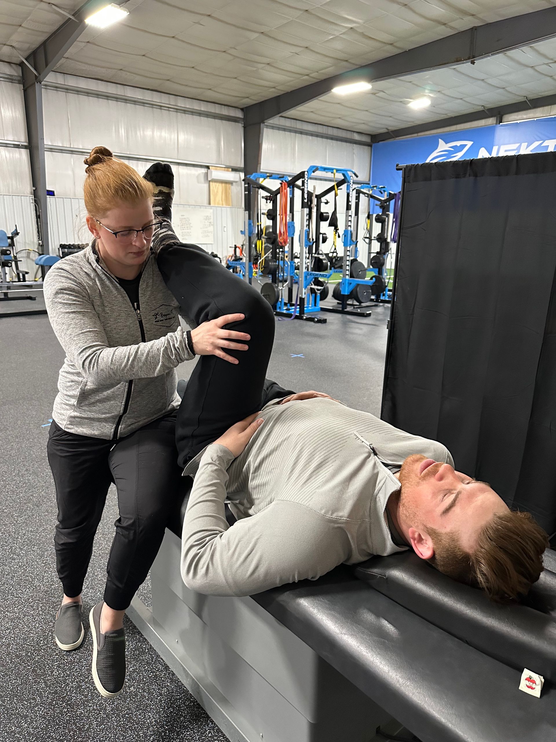 A person in a gray shirt lies on a table, leg raised. Another person in a gray jacket assists in a gym.
