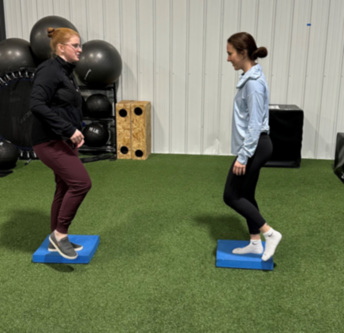 Two people balancing on blue platforms in a gym, facing each other.