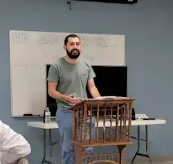 a man with a beard is standing at a podium in front of a whiteboard .