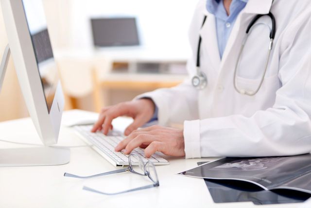 Médico tecleando en un escritorio con estetoscopio y gafas en una oficina luminosa.