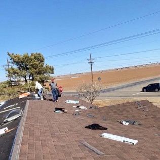 Damaged roof with scattered debris; several people inspect the edge near a road under a clear blue sky.