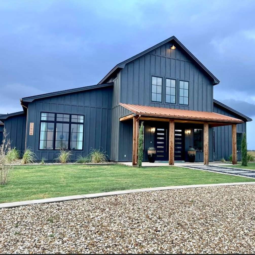 Modern dark blue farmhouse-style house with a front porch, large windows, and a gravel driveway at dusk
