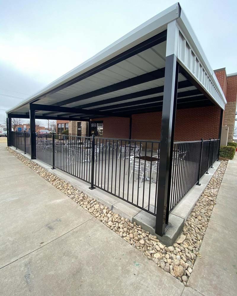 Covered outdoor patio with black metal fence, gray roof, and brick building beside a sidewalk.