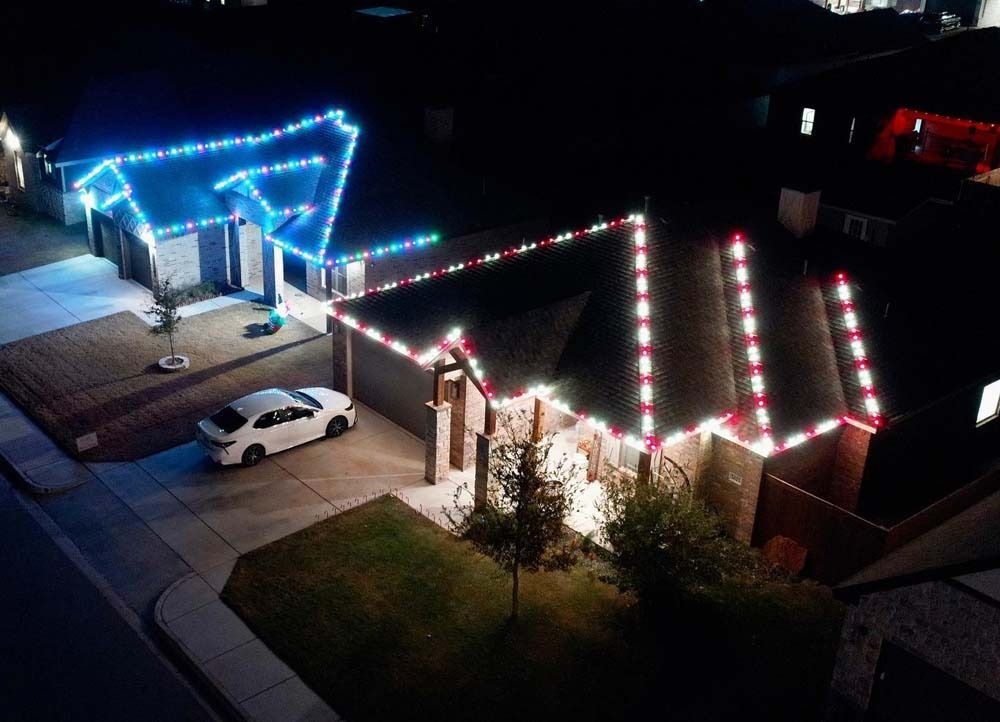 Nighttime aerial view of a house decorated with blue and red holiday lights, with a white car in the driveway