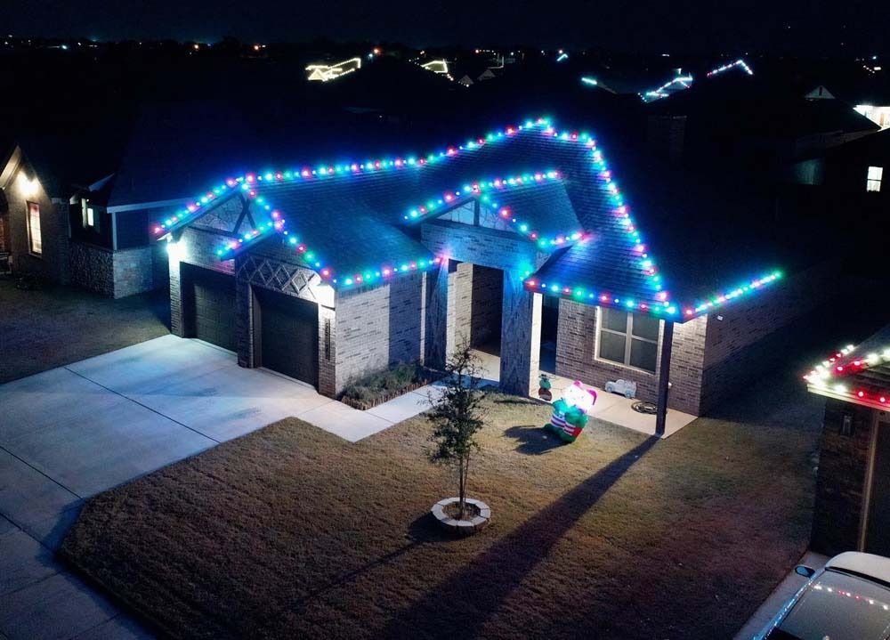 Single-story house at night outlined in blue and white holiday lights, with a lit front yard and driveway.