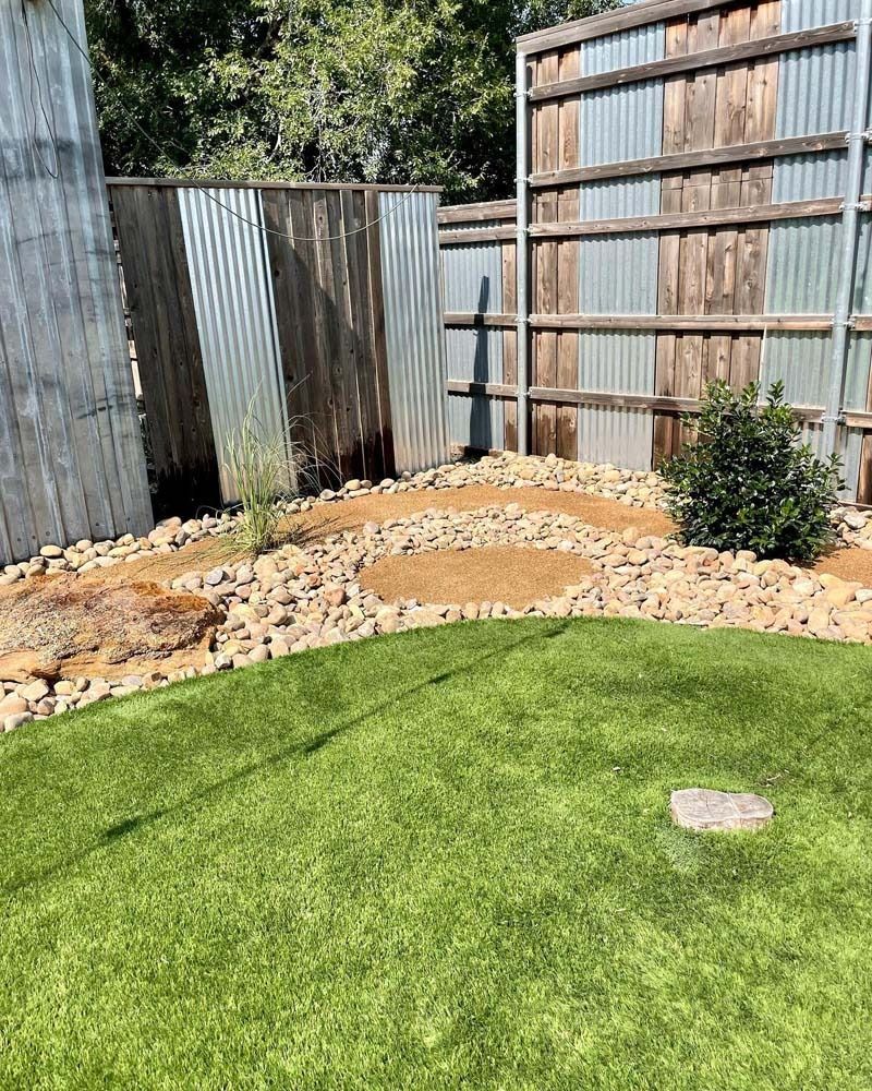 Backyard with green lawn, gravel border, corrugated metal fence, and a curved stone path.