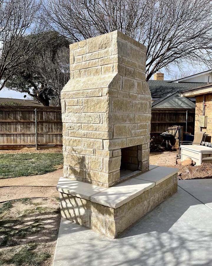 Stone outdoor fireplace on a concrete patio in a backyard, with wooden fence and trees behind it
