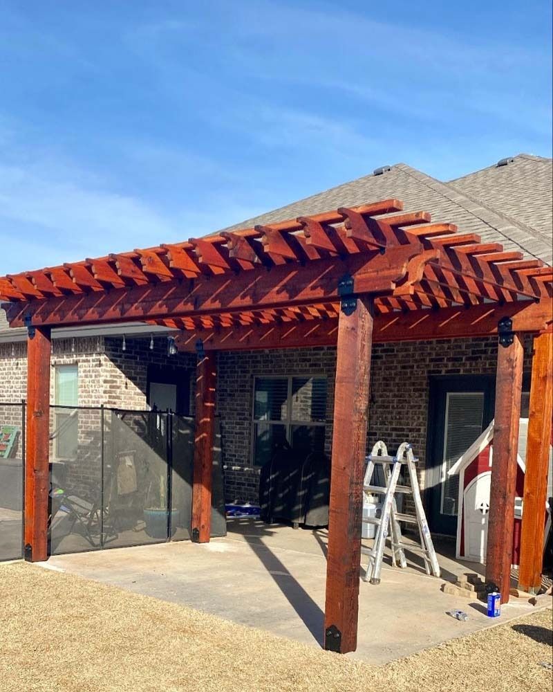Red wooden pergola over a patio with ladders and tools in a backyard, beside a stone wall and house.