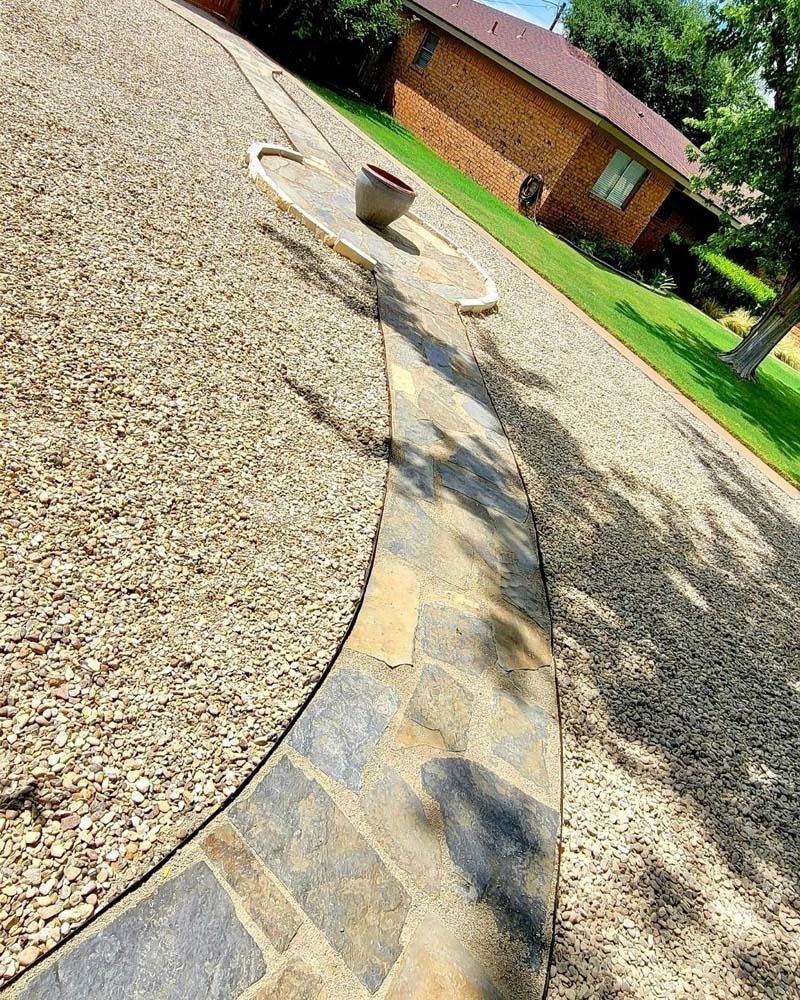 Curved stone border separating gravel and lawn in a sunny yard, with a small planter on the path.