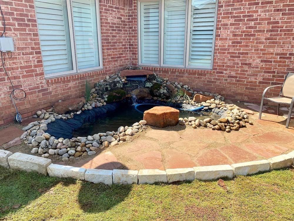 Backyard pond with rocks, plants, and a small round stump beside a brick house