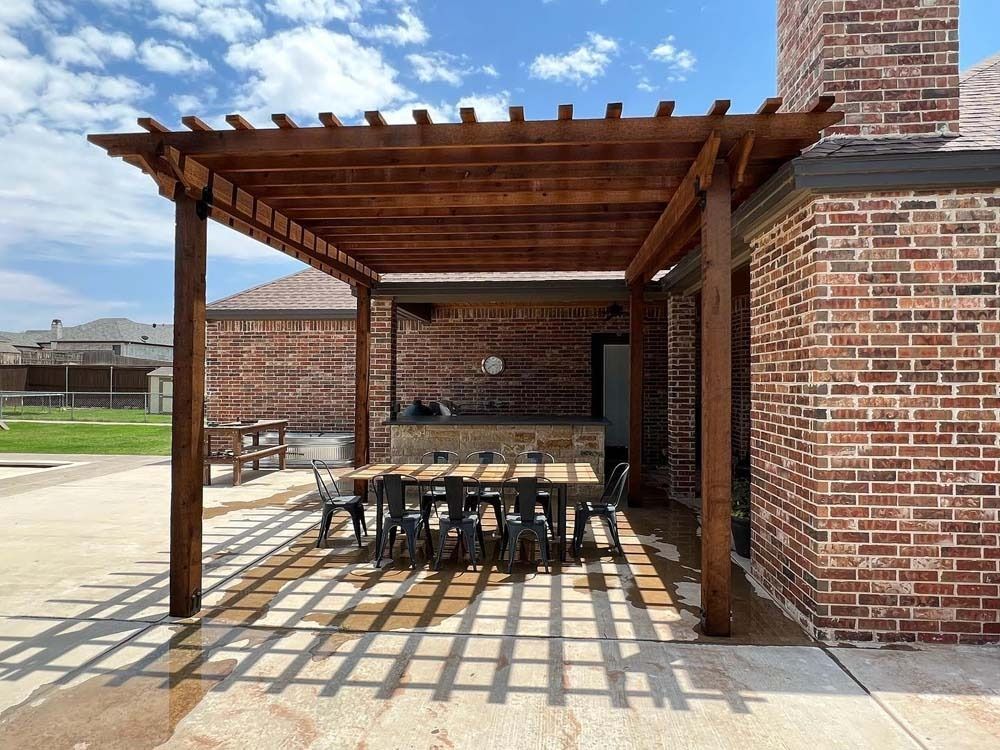Brick patio with wooden pergola and dining table beside a brick house under a blue sky.