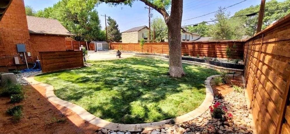 Sunny backyard with green lawn, brick walls, mature trees, and a curved stone border around the lawn