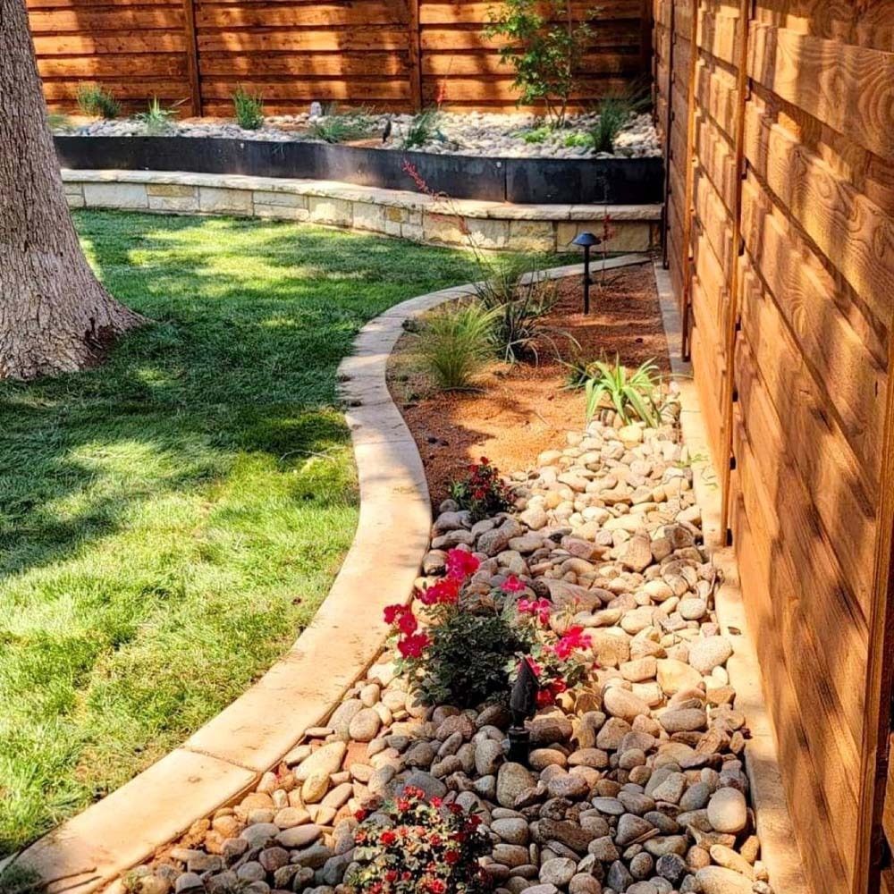 Curved garden bed with river rocks, red flowers, grass, and a wooden fence in sunlight