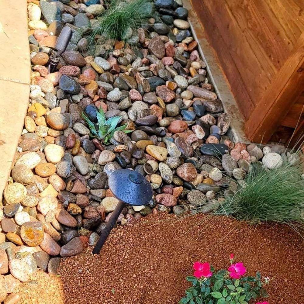 Garden bed with pebbles, a small black trowel, and pink flowers beside a wooden border.