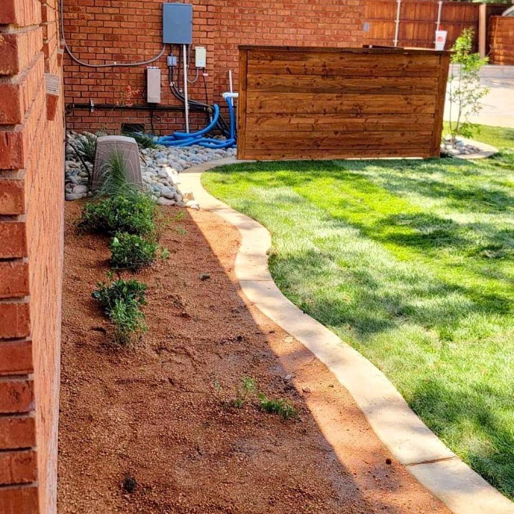 Freshly landscaped yard with red mulch bed, small shrubs, and a curved stone border beside green lawn and brick wall