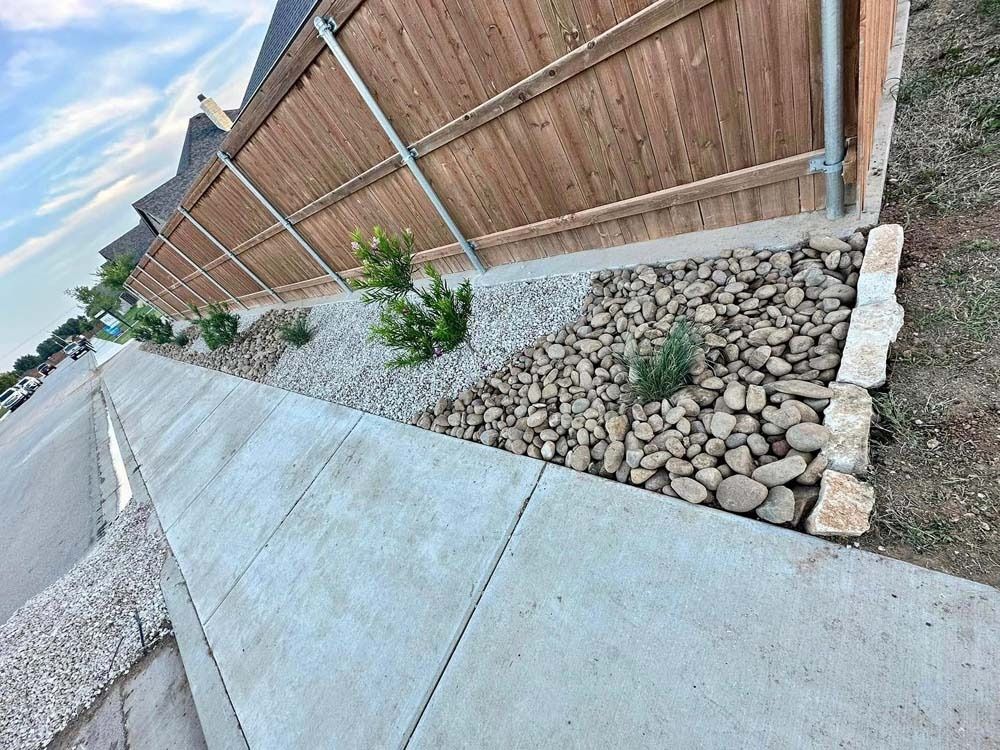 Concrete sidewalk beside a wooden fence and gravel drainage bed with small shrubs