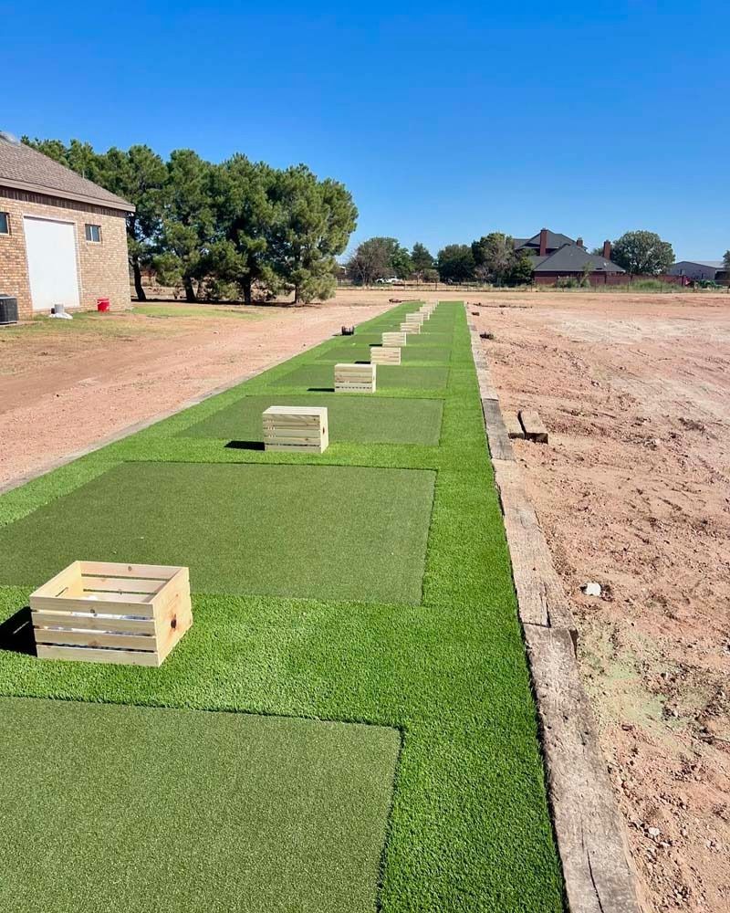 Long green turf strip with wooden crates beside a dirt lot near houses under a clear blue sky