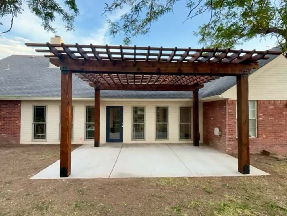 Covered patio with wooden pergola and concrete slab in front of a brick house