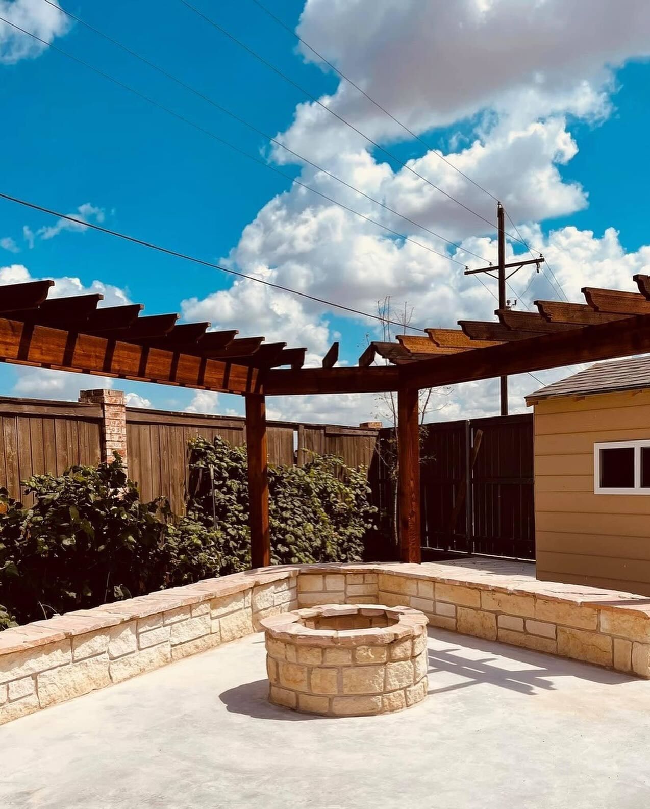 Backyard patio with a stone fire pit, wooden pergola, and tan shed under a blue sky.