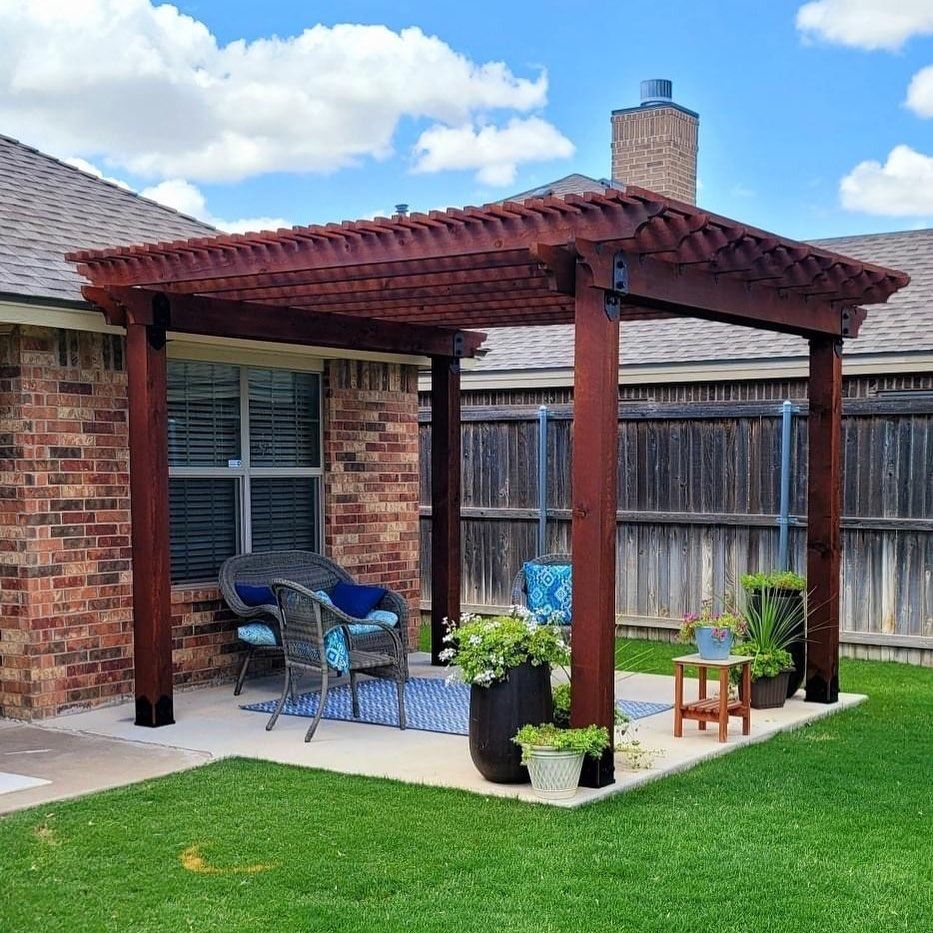 Brick patio with red pergola, chairs, potted plants, and grill in a fenced backyard.