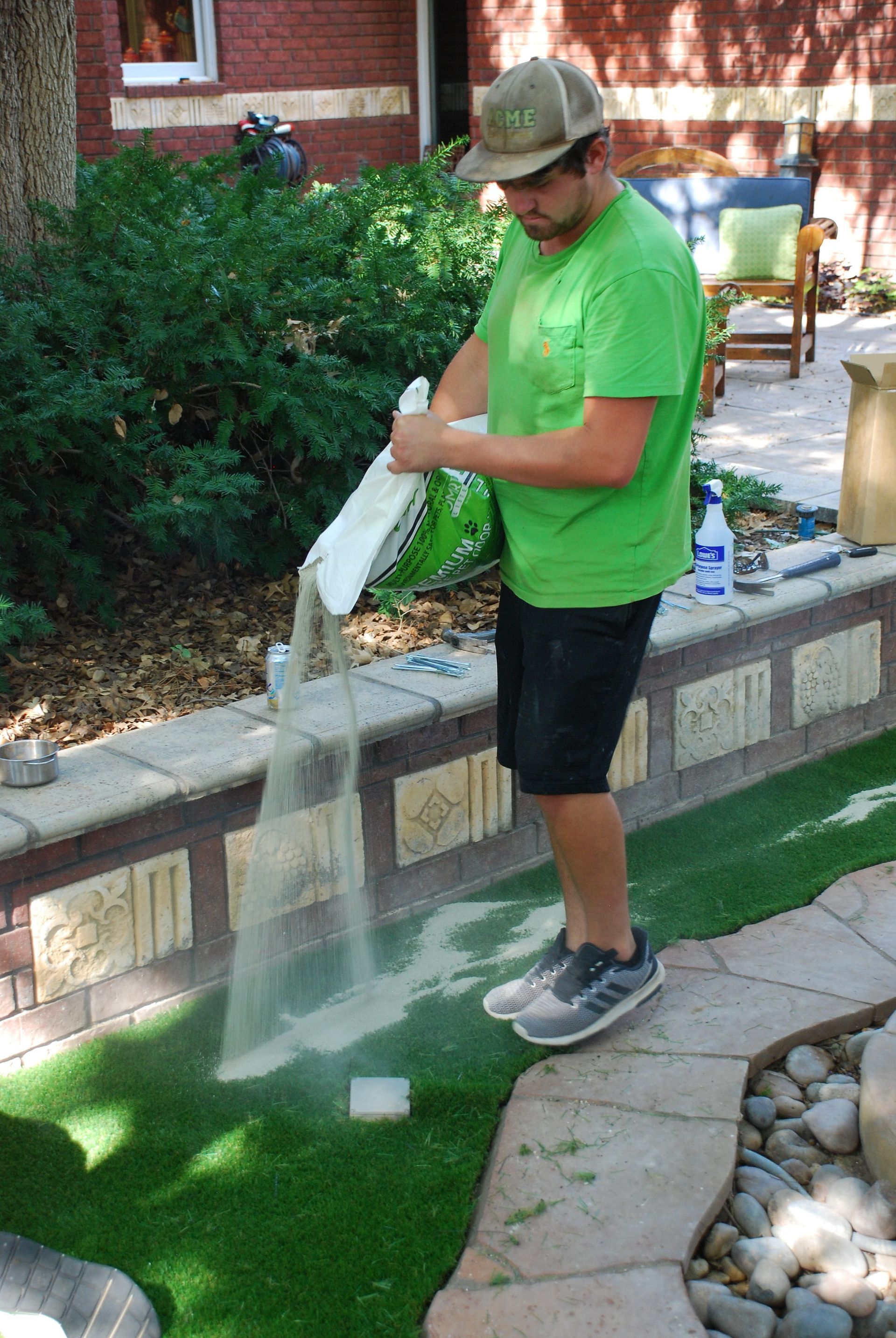 Man in green shirt and cap pouring water from a bag onto artificial turf in a backyard